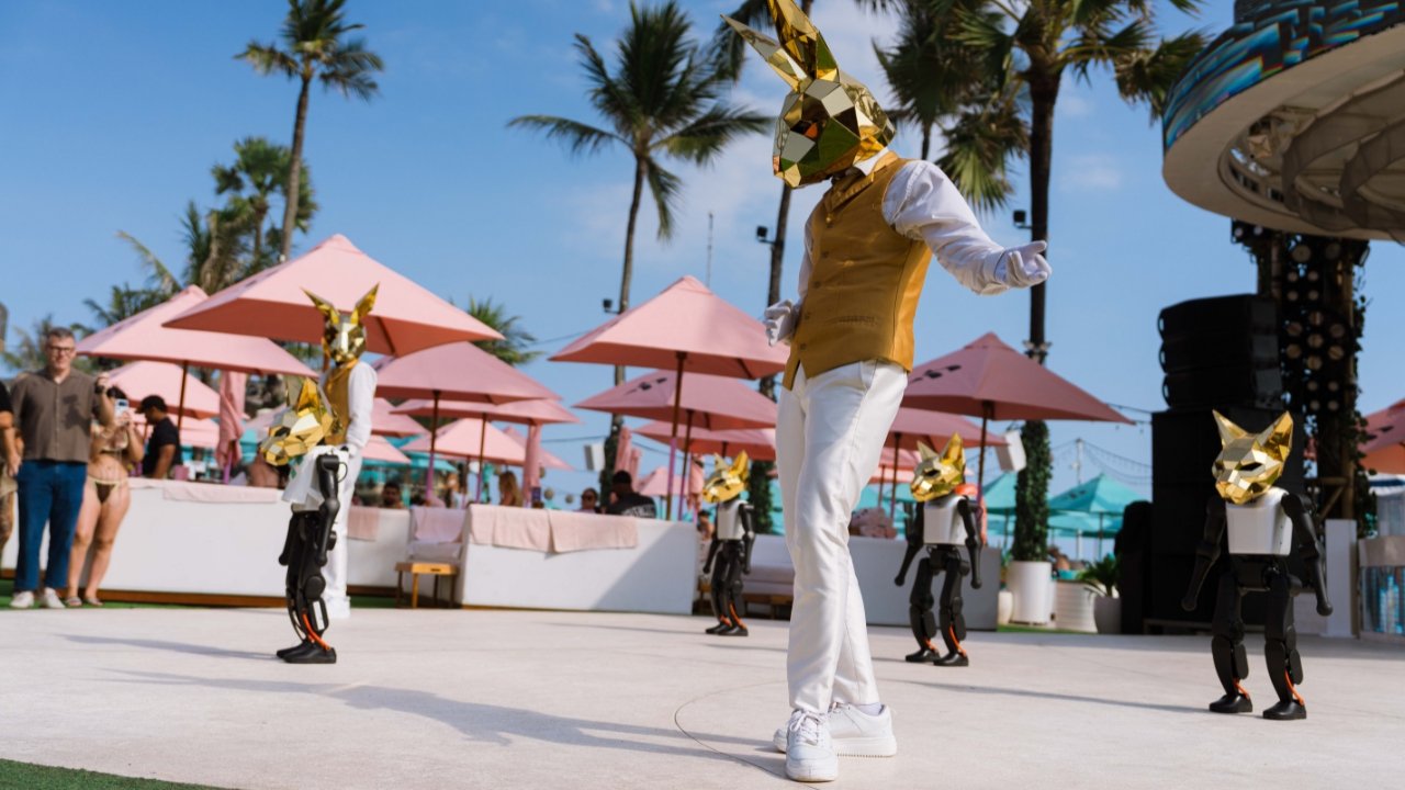 A performer in a gold rabbit mask leads four humanoid robots in a dance routine on the Finns Beach Club deck